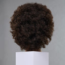 A dark brown curly wig displayed on a mannequin head, viewed from the back, resting on a white display block against a neutral gray background.