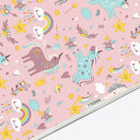 A close-up of a pink fabric cape printed with a whimsical unicorn pattern. The design features pastel pink unicorns, yellow stars, rainbows, clouds, and small decorative motifs.