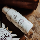 A bottle of Samson's Haircare Volumizing Dry Shampoo Matte Powder resting on a rustic wooden surface next to a brown leather bag, with a circular product container partially visible in the foreground.