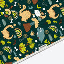 A close-up of a fabric or paper surface with a repeating whimsical dinosaur and nature print on a dark teal background. The pattern includes cartoon dinosaurs, trees, flowers, bones, and rainbows.