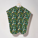 A baby bib or cape with a forest-themed pattern, hanging on a wooden hanger against a plain light gray wall.