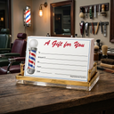 A barbershop-themed gift card holder and gift card set on a wooden counter inside a barbershop. The clear acrylic stand with gold-toned metal trim holds a white card printed with 'A Gift for You' in red script, a classic red, white, and blue barber pole graphic, and gift form fields.
