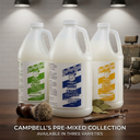 Three large white plastic jugs of Campbell's Pre-Mixed Shave Cream are displayed on a wooden table with grooming accessories, set against a blurred bathroom background with overhead lights. Text at the bottom reads 'CAMPBELL'S PRE-MIXED COLLECTION AVAILABLE IN THREE VARIETIES'.